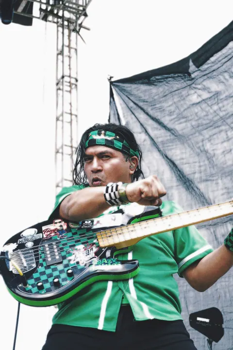 Bajista con vestimenta verde frente al escenario al aire libre.